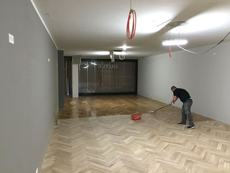 Ein Handwerker versiegelt einen geschliffenen Parkettboden in einem langen Raum